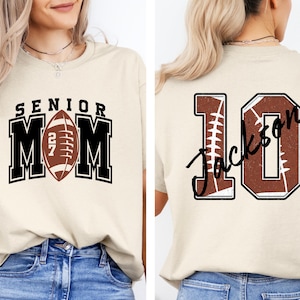 May include: A beige t-shirt featuring a front graphic with the text SENIOR MOM and a football icon with the number 27. The back graphic displays the number 10 with a football texture and the name Jackson written in black script across the center.