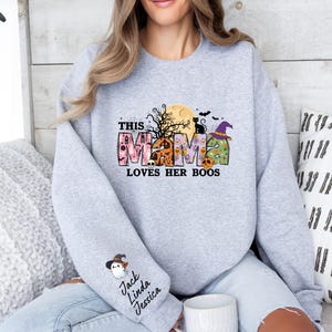 May include: Gray sweatshirt with a Halloween-themed design that says "This Mama Loves Her Boos". The design features a moon, bats, a black cat, a witch's hat, and a ghost. The sweatshirt has a small pocket on the left sleeve with the names "Jack, Linda, Jessica" written on it.
