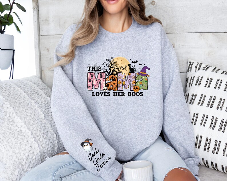 May include: Gray sweatshirt with a Halloween-themed design that says "This Mama Loves Her Boos". The design features a moon, bats, a black cat, a witch's hat, and a ghost. The sweatshirt has a small pocket on the left sleeve with the names "Jack, Linda, Jessica" written on it.