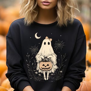 May include: Black sweatshirt with a white ghost holding a jack-o'-lantern. The ghost is wearing a bow and has a spooky, cute expression. The design is surrounded by stars, spiderwebs, and foliage.
