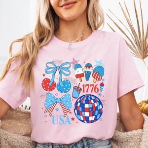 Puede incluir: Camiseta rosa claro con un dise&ntilde;o patri&oacute;tico. El gr&aacute;fico incluye lazos, cerezas, helados y una bola de discoteca en colores rojo, blanco y azul. Tambi&eacute;n aparece el texto "1776" y "USA".