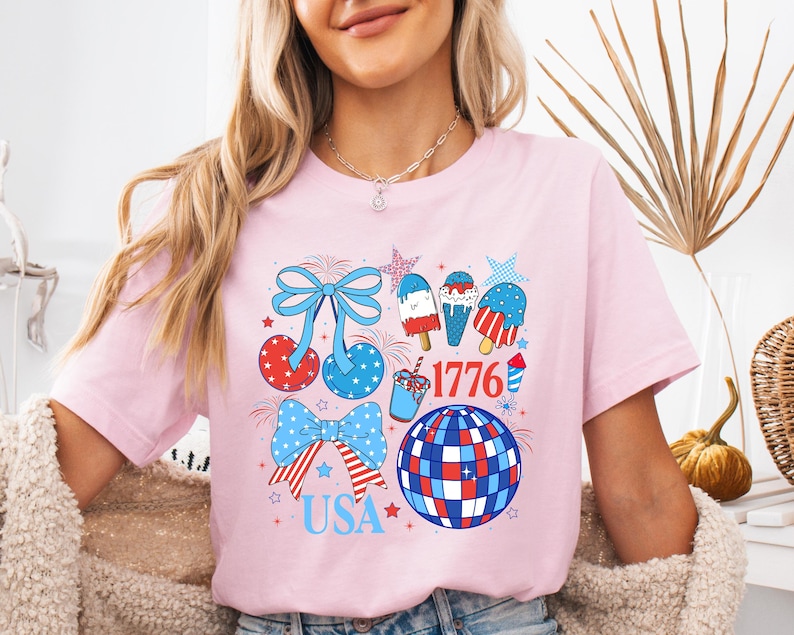Puede incluir: Camiseta rosa claro con un dise&ntilde;o patri&oacute;tico. El gr&aacute;fico incluye lazos, cerezas, helados y una bola de discoteca en colores rojo, blanco y azul. Tambi&eacute;n aparece el texto "1776" y "USA".