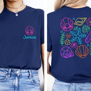 May include: Navy blue t-shirt with a seashell design and the name "Jessica" in teal. The back of the shirt features a colorful neon design of seashells, starfish, and flowers. A casual, beach-themed tee.