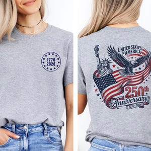 Pode incluir: Camiseta cinza mesclada com design patri&oacute;tico. A frente apresenta um c&iacute;rculo azul marinho com o texto "1776 2026". A parte de tr&aacute;s mostra a Est&aacute;tua da Liberdade, uma &aacute;guia e a bandeira americana, com texto "250th Anniversary".