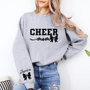 May include: Gray sweatshirt with the text "CHEER MOM" and a black bow. The left sleeve has the text "Jessica" and a black bow.