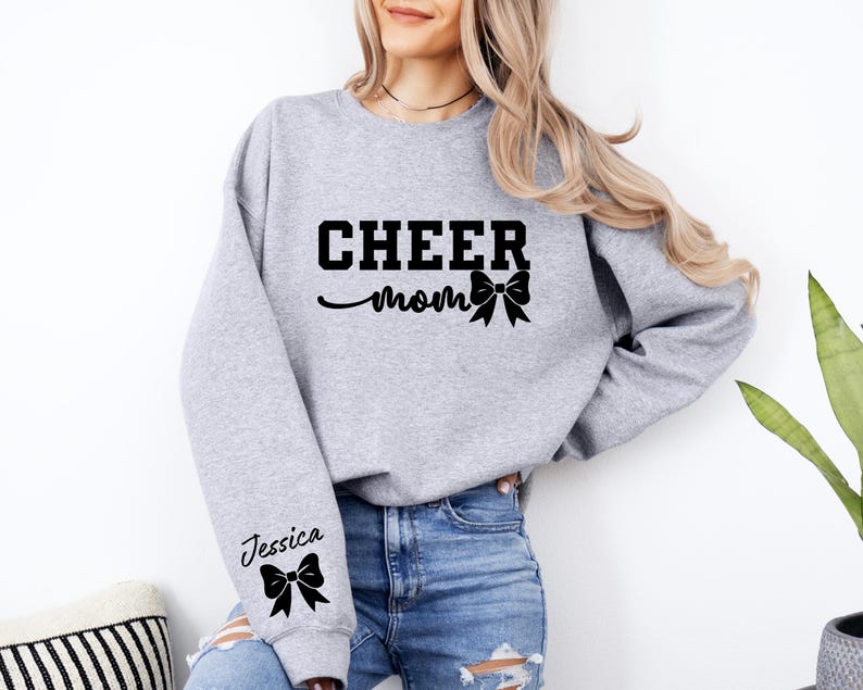 May include: Gray sweatshirt with the text "CHEER MOM" and a black bow. The left sleeve has the text "Jessica" and a black bow.