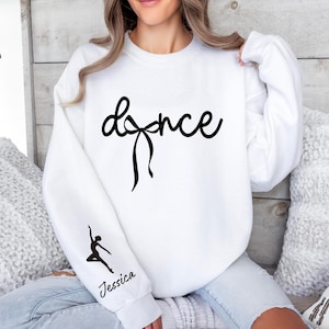 May include: White sweatshirt with the word "dance" in a cursive font, featuring a bow design. A dancer silhouette and the name "Jessica" are printed on the sleeve. The sweatshirt is a casual garment, suitable for dance enthusiasts.