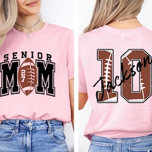May include: A light pink t-shirt featuring a front graphic with the text SENIOR MOM and a football icon with the number 27. The back of the shirt displays the number 10 with a football texture and the name Jackson written in black script.