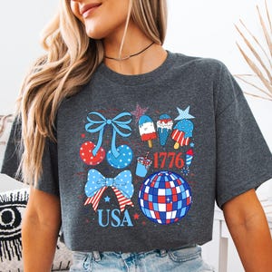 Puede incluir: Camiseta gris oscuro con un dise&ntilde;o patri&oacute;tico con lazos, helados y una bola de discoteca en rojo, blanco y azul. La palabra "USA" y el n&uacute;mero "1776" tambi&eacute;n est&aacute;n incluidos en el dise&ntilde;o.