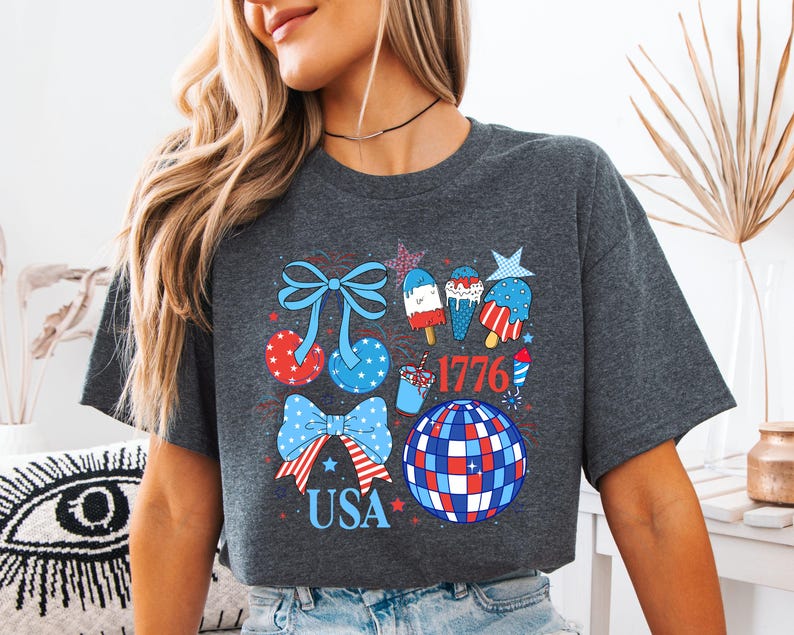 Puede incluir: Camiseta gris oscuro con un dise&ntilde;o patri&oacute;tico con lazos, helados y una bola de discoteca en rojo, blanco y azul. La palabra "USA" y el n&uacute;mero "1776" tambi&eacute;n est&aacute;n incluidos en el dise&ntilde;o.