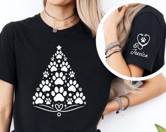 Könnte beinhalten: Schwarzes T-Shirt mit einem weißen Weihnachtsbaum-Design aus Pfotenabdrücken. Das Shirt hat außerdem ein weißes Stethoskop mit einem Pfotenabdruck in der Mitte und den Namen "Jessica" auf dem Ärmel.