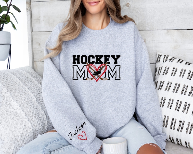 Pu&ograve; includere: Felpa grigia con il testo "HOCKEY MOM" in nero con un cuore rosso e bastoni da hockey incrociati. Il testo "Jackson" &egrave; sulla manica con un cuore rosso.