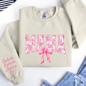 May include: A beige sweatshirt with the word "MAMA" printed in pink floral letters with a pink bow. The left sleeve has the names "Calvin, Karissa, Jessica" written in pink cursive with a pink heart.