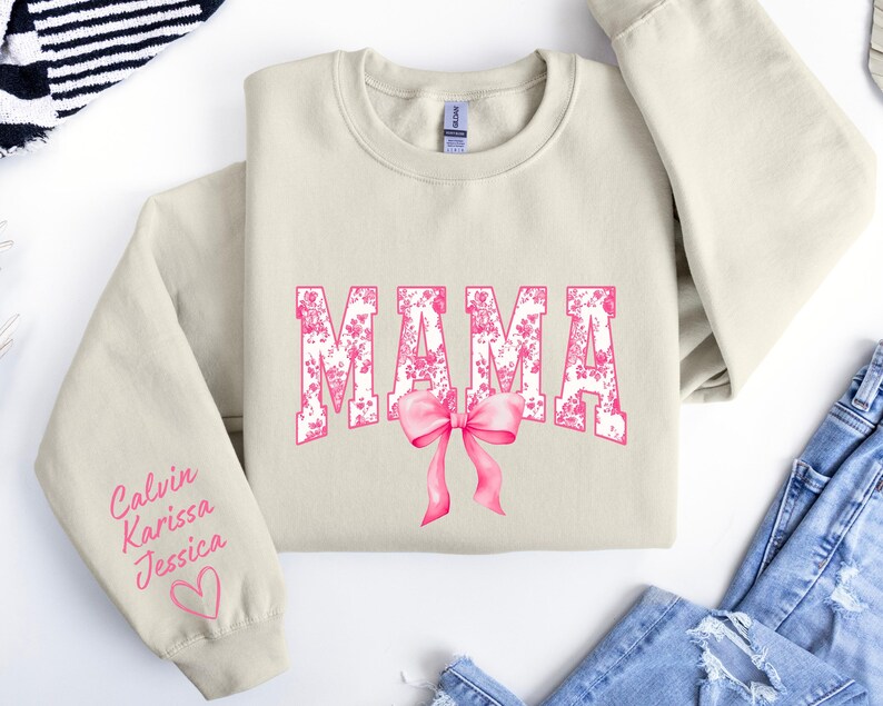 May include: A beige sweatshirt with the word "MAMA" printed in pink floral letters with a pink bow. The left sleeve has the names "Calvin, Karissa, Jessica" written in pink cursive with a pink heart.