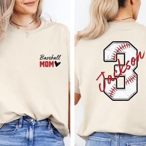 May include: A beige t-shirt with the text "Baseball Mom" and a small red heart on the front. The back of the shirt has a large white number 3 with a red baseball stitched design and the text "Jackson" in red cursive writing.