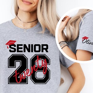 May include: A light gray t-shirt with the word "SENIOR" above the number "20" in a black and red design. The sleeve has a small red graduation cap and the name "Olivia" in black script.