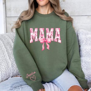 May include: A green sweatshirt with the word "MAMA" in pink and white floral print. The sweatshirt has a pink bow and the name "Jessica" written on the sleeve with a heart.