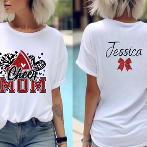Puede incluir: Camiseta blanca con un dise&ntilde;o rojo, negro y blanco que dice "Cheer Mom" en la parte delantera. La parte posterior de la camiseta tiene un lazo rojo y el nombre "Jessica" en negro.