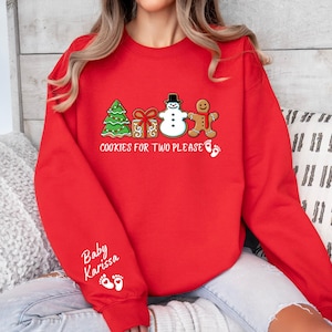 May include: A red sweatshirt with a festive design. The design includes a green Christmas tree, a wrapped gift, a snowman, and a gingerbread person. The text "COOKIES FOR TWO PLEASE" is printed below the images. The sleeve has the text "Baby Karissa" and footprints.