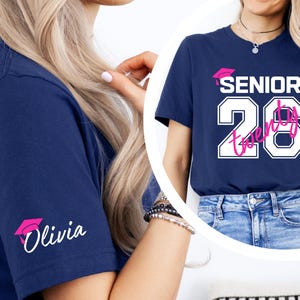 May include: Navy blue t-shirt with the text "SENIOR 2026" in white and pink, featuring a graduation cap graphic. The sleeve displays a pink graduation cap and the name "Olivia" in white. The shirt is a casual style.