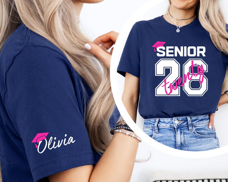 May include: Navy blue t-shirt with the text "SENIOR 2026" in white and pink, featuring a graduation cap graphic. The sleeve displays a pink graduation cap and the name "Olivia" in white. The shirt is a casual style.