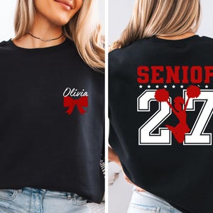 May include: A black sweatshirt featuring a red bow and the name Olivia on the front left chest. The back displays the word SENIOR in red, the number 27 in large white block font, and a red silhouette of a cheerleader holding pom-poms.