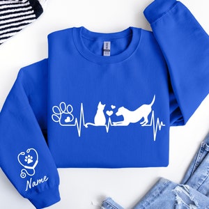 May include: A royal blue sweatshirt with a white design of a cat and dog with a heartbeat line and paw print. The sleeve has a white stethoscope with a paw print and the word "Name" underneath.