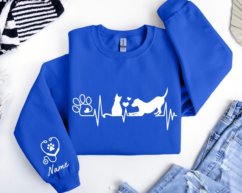 May include: A royal blue sweatshirt with a white design of a cat and dog with a heartbeat line and paw print. The sleeve has a white stethoscope with a paw print and the word "Name" underneath.