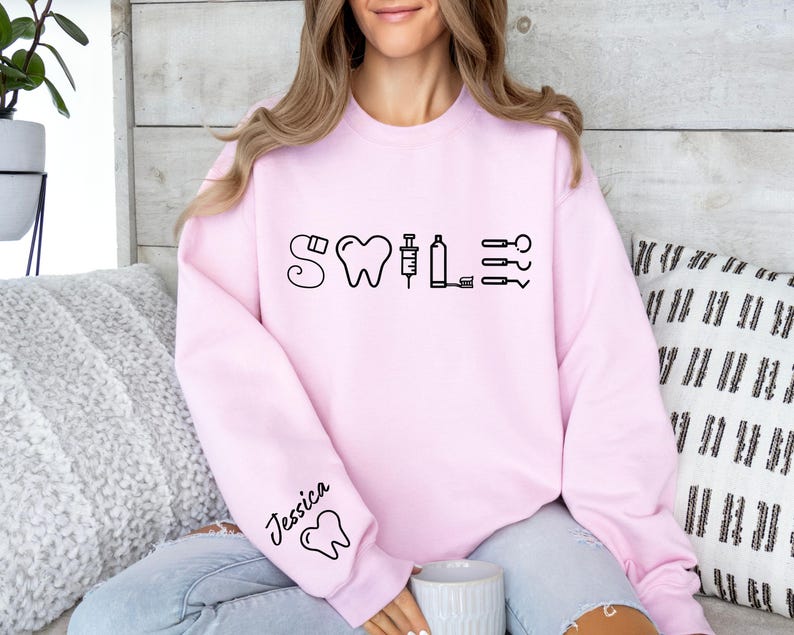 May include: Pink sweatshirt with a graphic of dental tools and the word "Smile" on the front. The sweatshirt has the name "Jessica" and a tooth graphic on the left sleeve.