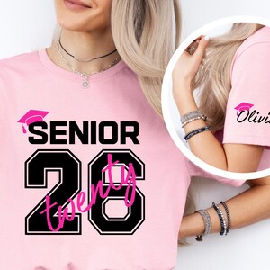 May include: Pink t-shirt with the word "SENIOR" above the number "26". The number is outlined in black with the word "twenty" in pink inside. A graduation cap graphic is above the word "SENIOR". The sleeve has a graduation cap graphic and the name "Olivia".