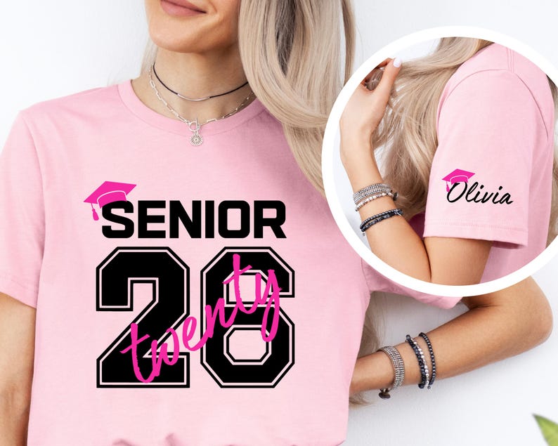 May include: Pink t-shirt with the word "SENIOR" above the number "26". The number is outlined in black with the word "twenty" in pink inside. A graduation cap graphic is above the word "SENIOR". The sleeve has a graduation cap graphic and the name "Olivia".