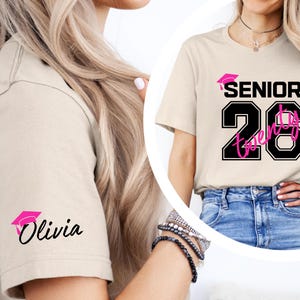 May include: Beige t-shirt with the text "SENIOR Twenty" in black and pink, with a graduation cap graphic. The sleeve features a pink graduation cap and the name "Olivia" in black script. The shirt is worn with blue jeans.