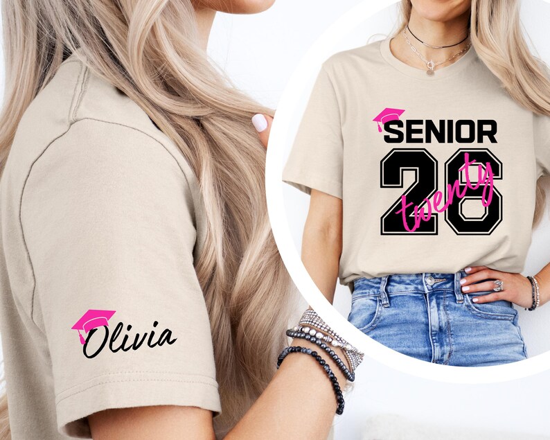 May include: Beige t-shirt with the text "SENIOR Twenty" in black and pink, with a graduation cap graphic. The sleeve features a pink graduation cap and the name "Olivia" in black script. The shirt is worn with blue jeans.