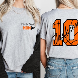 Peut inclure: T-shirt gris avec le texte "Basketball Mom" et un c&oelig;ur noir sur le devant. Le dos du t-shirt a un grand ballon de basket orange avec le num&eacute;ro 10 et le nom "Jackson" &eacute;crit &agrave; l'int&eacute;rieur.