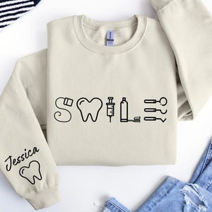 May include: A beige sweatshirt with a black graphic of dental tools spelling out the word "SMILE". The left sleeve has the name "Jessica" and a tooth graphic.