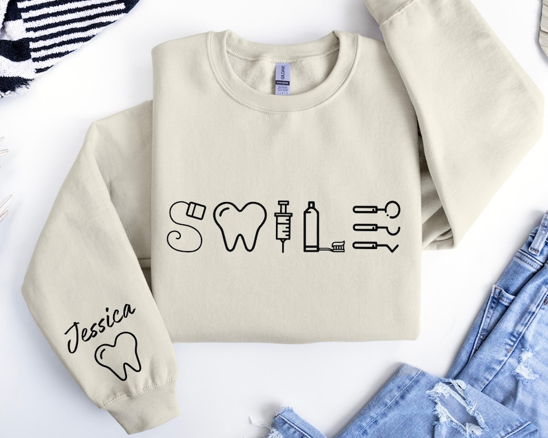 May include: A beige sweatshirt with a black graphic of dental tools spelling out the word "SMILE". The left sleeve has the name "Jessica" and a tooth graphic.