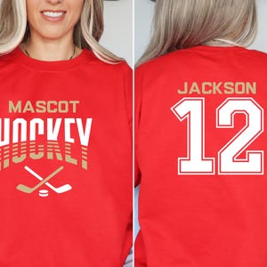 K&ouml;nnte beinhalten: Roter Pullover mit einem gold-wei&szlig;en Hockey-Design auf der Vorderseite. Die R&uuml;ckseite des Pullovers hat eine wei&szlig;e Nummer 12 mit dem Namen "JACKSON" dar&uuml;ber.