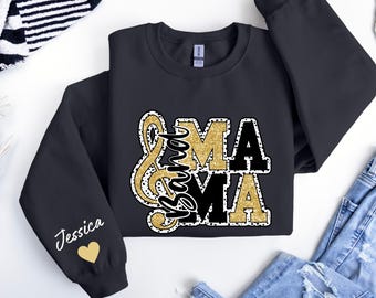 Può includere: Felpa nera con la scritta "Band Mama" in un motivo a forma di nota musicale, e la parola "MAMA" in glitter dorati e lettere nere. La manica ha il nome "Jessica" e un cuore dorato.