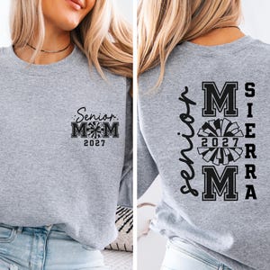 May include: A heather gray sweatshirt with black text and a pom-pom graphic. The front of the sweatshirt reads "Senior Mom 2027." The back of the sweatshirt says "Senior Sierra Mom 2027" in a vertical layout.