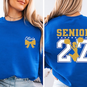 May include: A royal blue crewneck sweatshirt featuring a gold bow and the name Olivia on the front left chest. The back displays the word SENIOR in gold above the number 27 in white, with a gold silhouette of a cheerleader holding pom-poms.