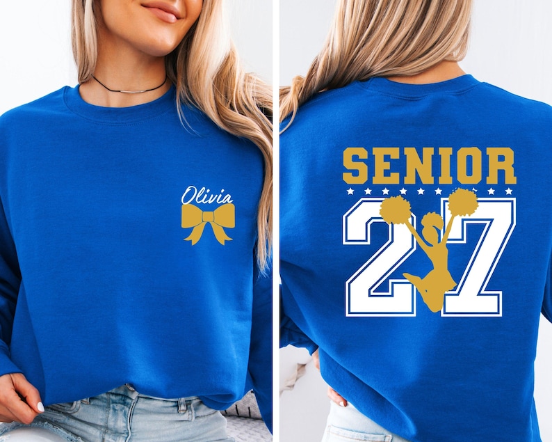 May include: A royal blue crewneck sweatshirt featuring a gold bow and the name Olivia on the front left chest. The back displays the word SENIOR in gold above the number 27 in white, with a gold silhouette of a cheerleader holding pom-poms.