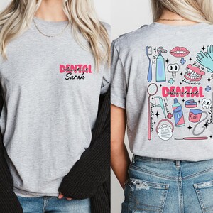 May include: Grey t-shirt with a pink "Dental Assistant Sarah" text on the front and a colourful illustration of dental tools and equipment on the back.