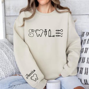 May include: A beige sweatshirt with a black graphic of a tooth, syringe, and other dental tools with the word "SMILE" above it. The text "Jessica" is written on the sleeve of the sweatshirt.