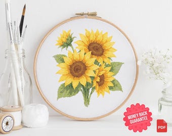 May include: Cross stitch pattern featuring a bouquet of three yellow sunflowers with green leaves. The pattern is displayed in a wooden embroidery hoop.