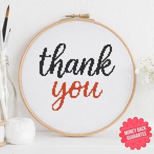Może przedstawiać: Okrągła tamborkowa z napisem "thank you" wyszytym czarną i pomarańczową nicią na białym materiale. Tamborek jest wykonany z drewna i jest prezentowany na białej powierzchni. Obraz zawiera czerwoną naklejkę "Money Back Guarantee".
