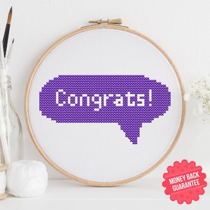May include: A cross-stitch embroidery in a wooden hoop features a purple speech bubble with the word "Congrats!" in white. The embroidery is set against a white background, with a few art supplies and flowers in the background.