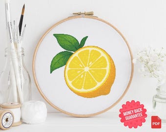 May include: A cross-stitch embroidery of a lemon with green leaves, set in a wooden hoop. The lemon is yellow with a white center, and the leaves are green. The background is white. A red "Money Back Guarantee" sticker is in the lower right corner.