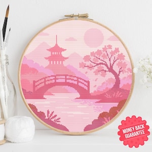 May include: A pink cross-stitch embroidery of a Japanese landscape, featuring a pagoda, bridge, and cherry blossom tree. The artwork is displayed in a wooden embroidery hoop. The scene is rendered in shades of pink and red.