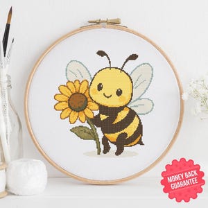May include: A cross-stitch embroidery hoop featuring a cartoon bee holding a sunflower. The bee is yellow and black with light blue wings. The sunflower is yellow and brown. The hoop is set against a white background.
