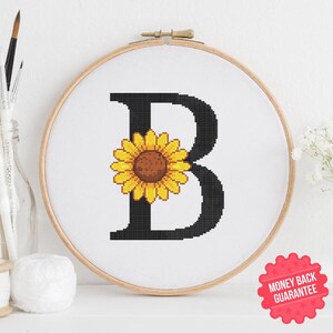May include: Cross-stitch embroidery of the letter "B" with a sunflower in the center, set in a wooden hoop. The letter is black, the sunflower is yellow and brown, and the background is white. A "Money Back Guarantee" sticker is visible.